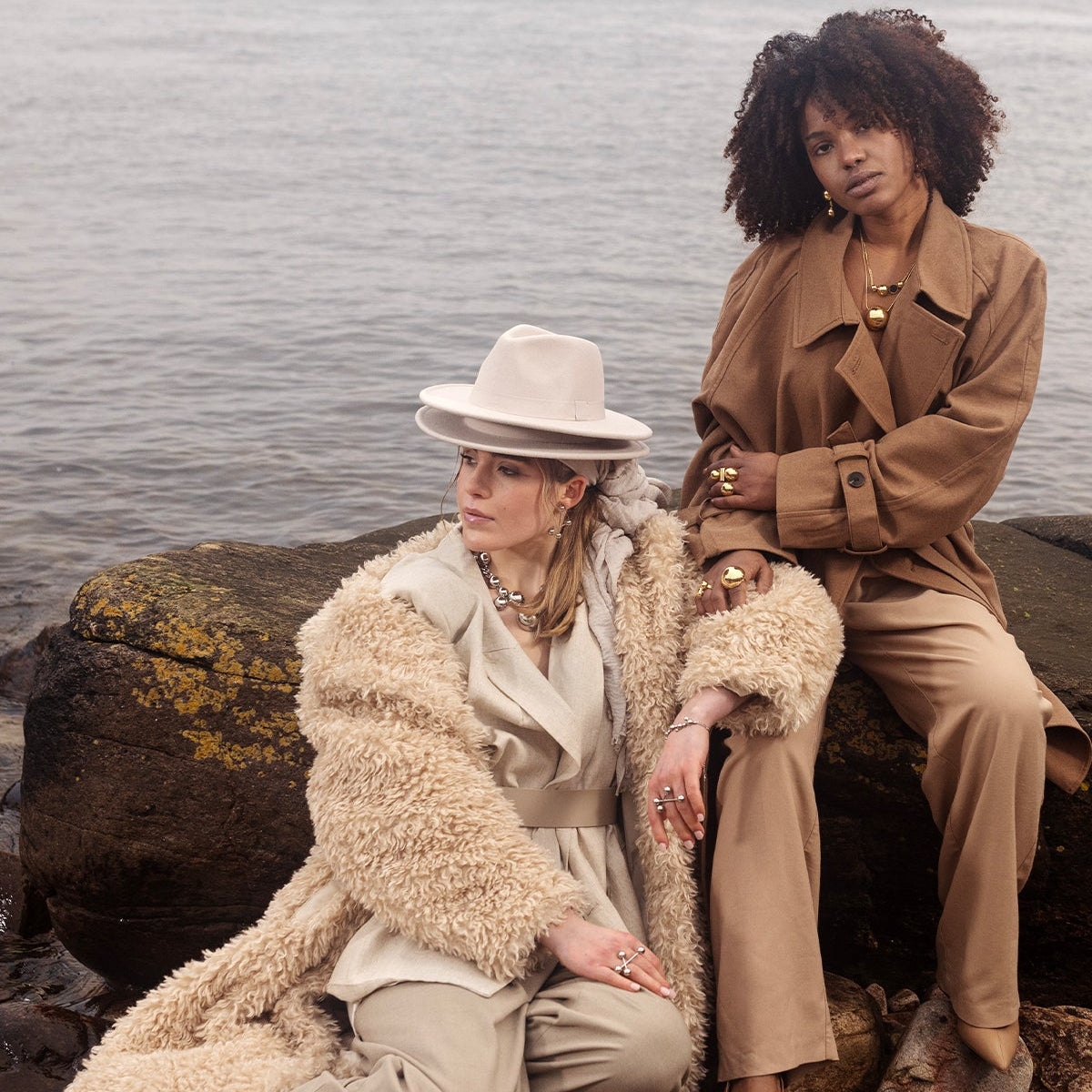Two women sitting on rocks by a body of water, wearing stylish outfits.