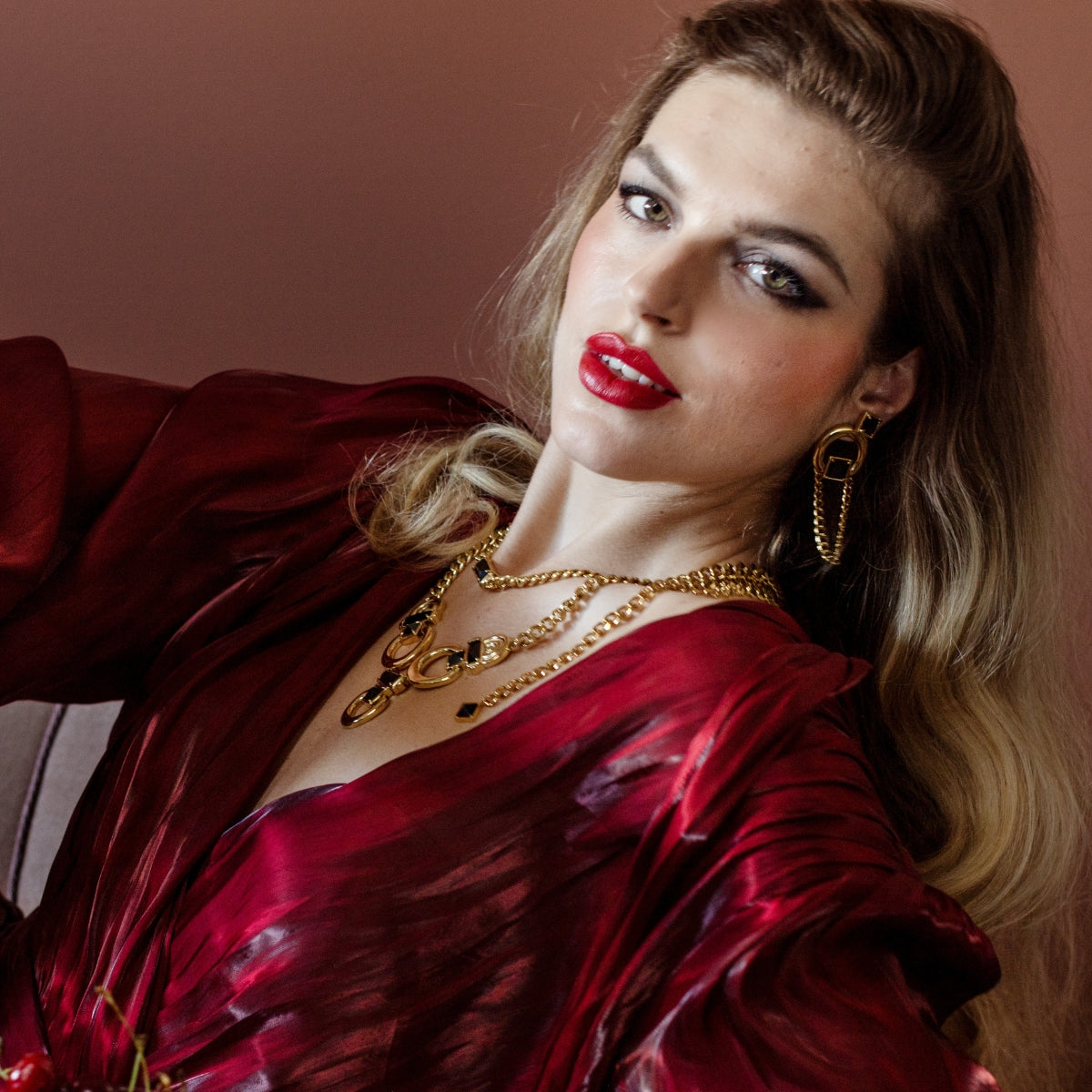 A woman with blonde hair wearing a shiny deep red satin robe and layered gold necklaces, posing against a warm brown background.