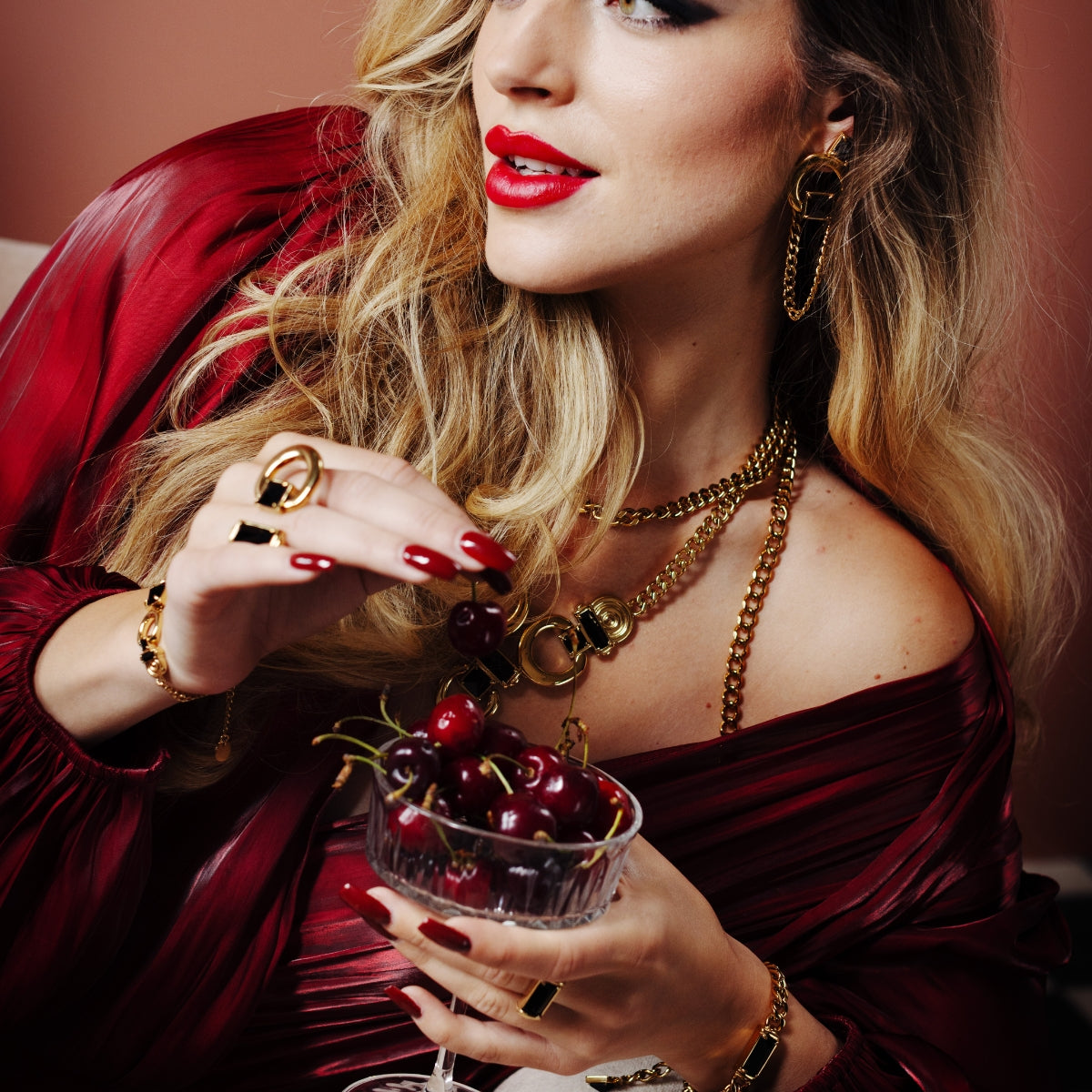A close-up lifestyle shot of a woman with blonde hair wearing a deep burgundy off-the-shoulder dress and multiple gold jewelry pieces, holding a glass of cherries.