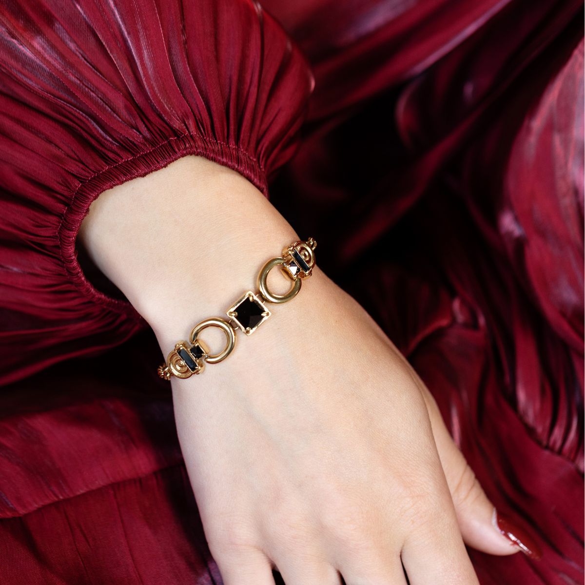 A gold-tone chain bracelet with black rectangular and circular links, worn on a woman's wrist. The background consists of a dark red pleated fabric, likely a dress or scarf.