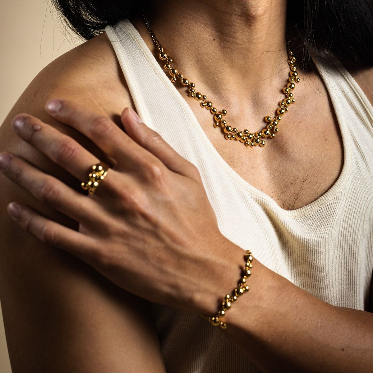 Person wearing gold necklace, ring, and bracelet with a neutral background