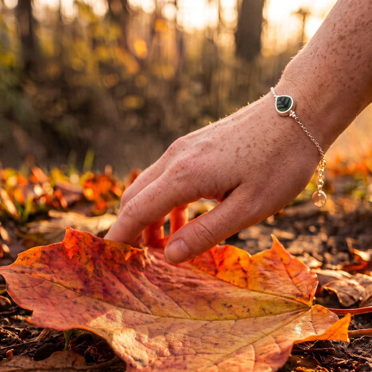 A close-up lifestyle shot of a person's hand touching a large orange autumn maple leaf on the ground, wearing a delicate silver chain bracelet with a green heart-shaped stone and a small dangling bead.