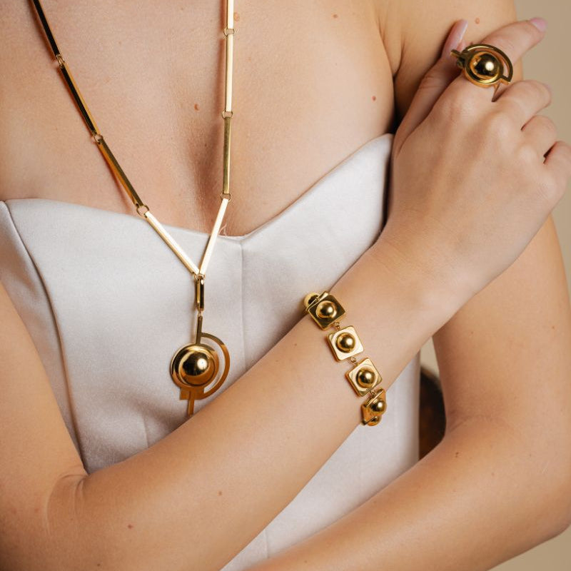 Gold necklace, ring, and bracelet on a person wearing a white top against a neutral background
