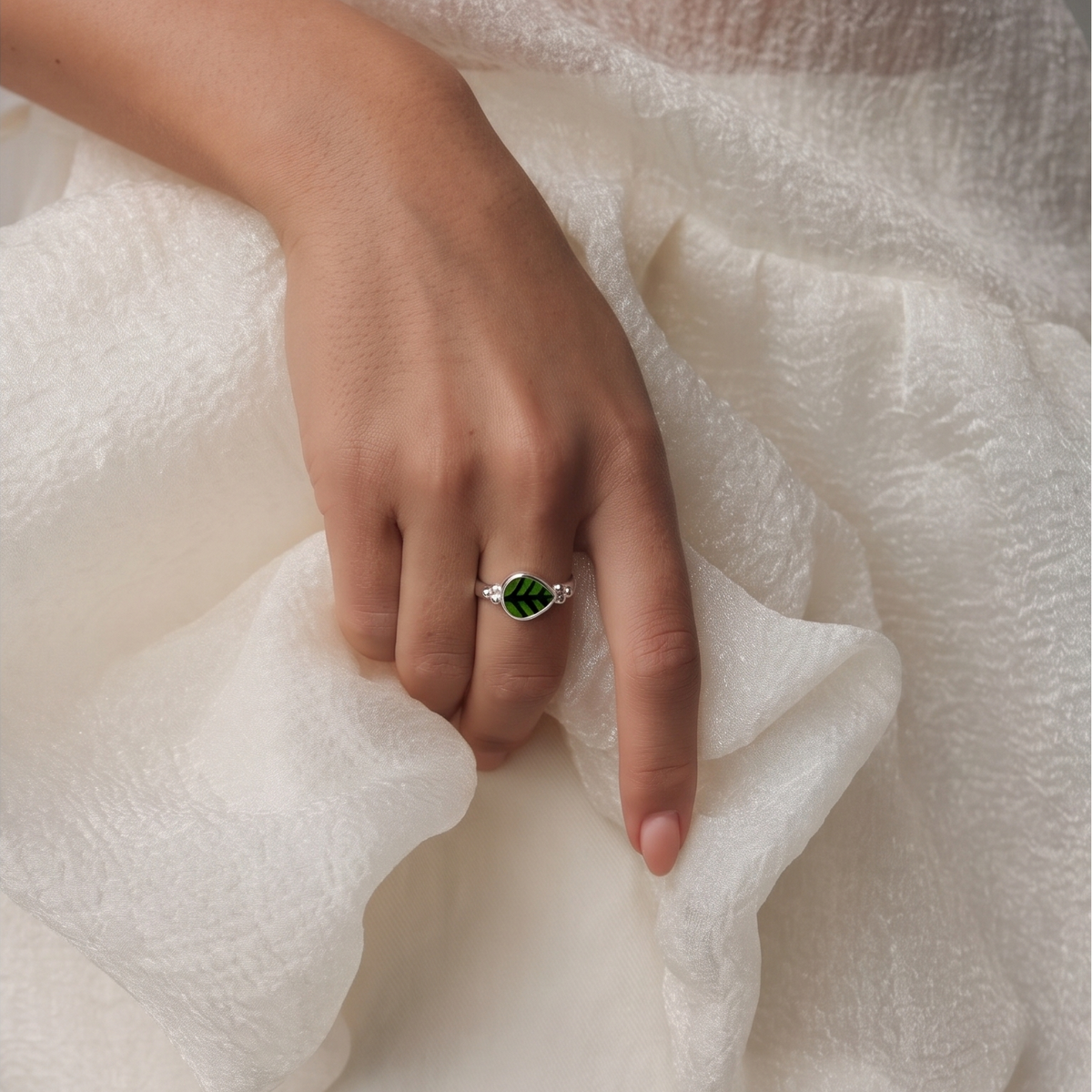 A close-up of a hand wearing a silver ring with a marquise-cut green gemstone, resting on soft, textured white fabric.