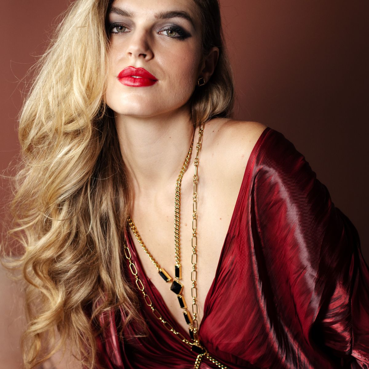 A close-up portrait of a woman with long wavy blonde hair, wearing a deep burgundy satin dress and layered gold necklaces, set against a solid dark brown background.
