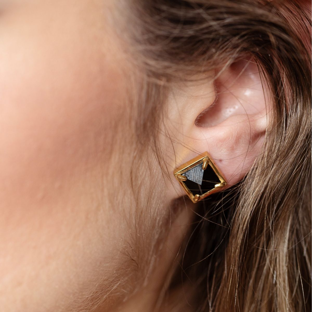 A close-up of a person's ear wearing a geometric stud earring with a faceted black center and a gold-tone border.