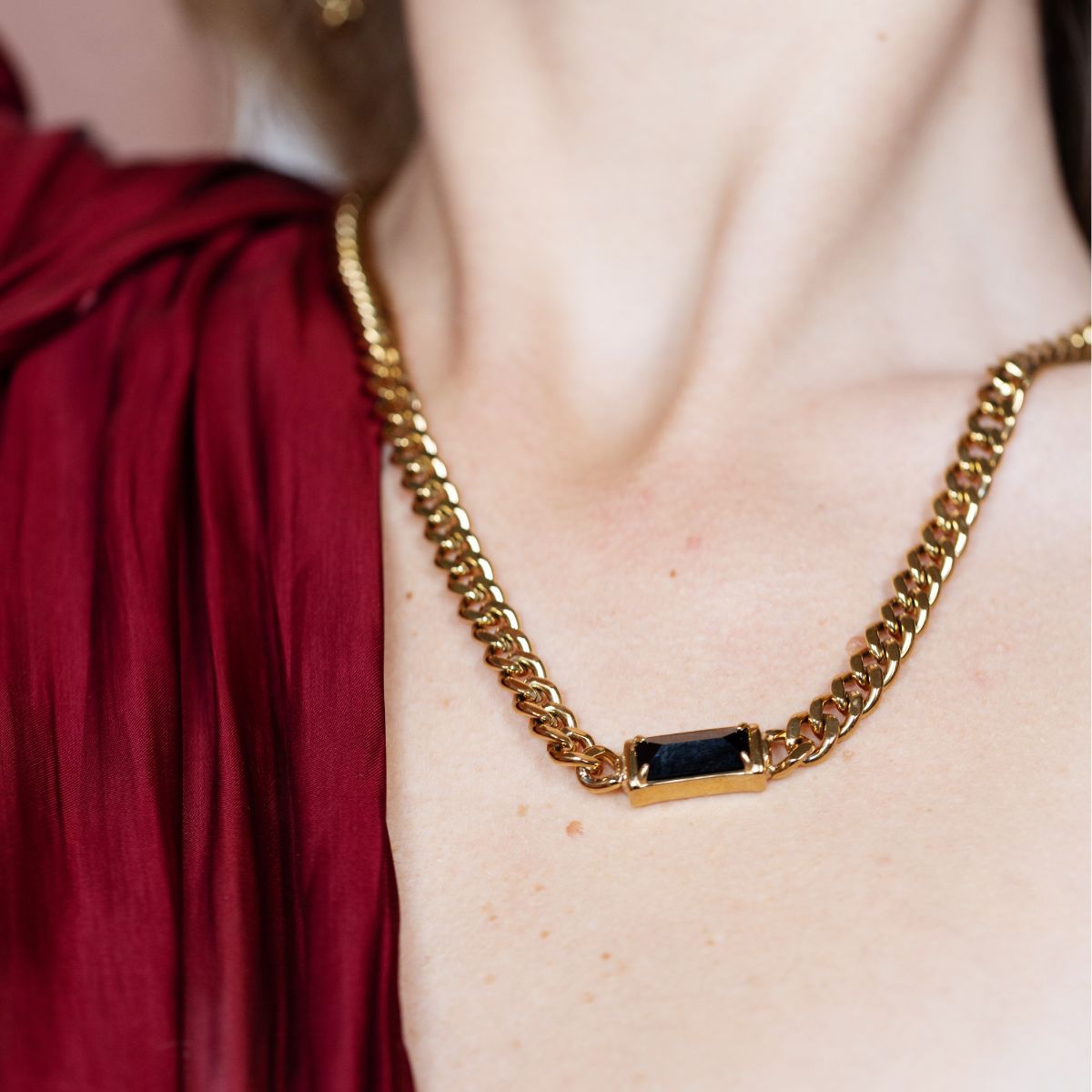 A close-up of a gold chain necklace with a rectangular black gemstone centerpiece, worn by a model in a red top.