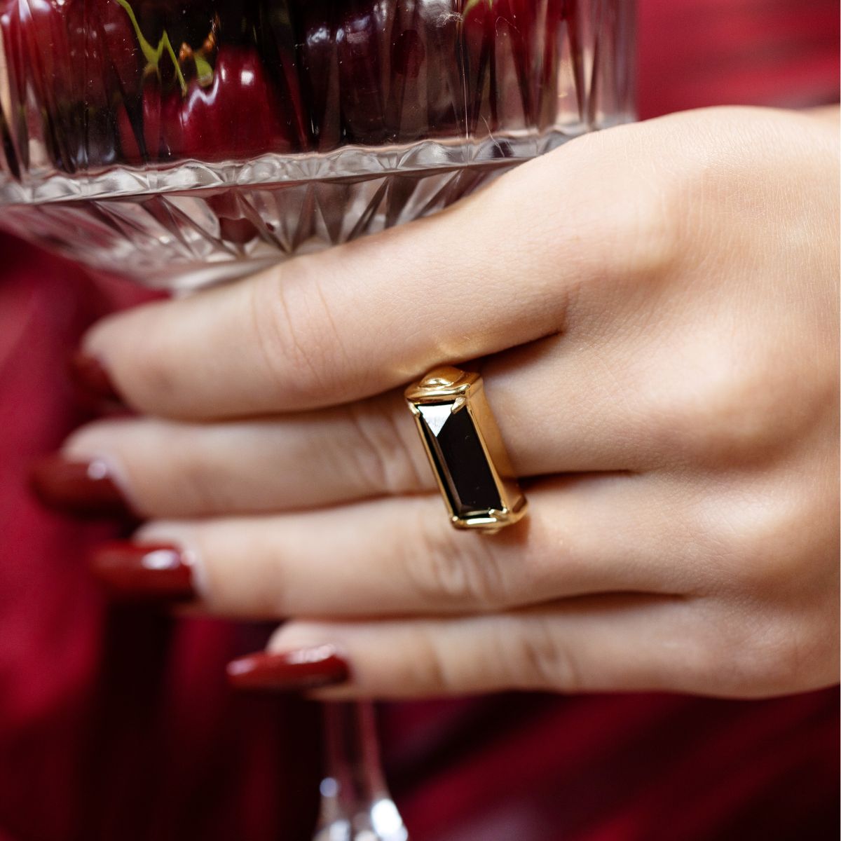 A close-up of a hand wearing a gold ring with a rectangular black inlay, holding a faceted glass of dark red liquid against a red fabric background.