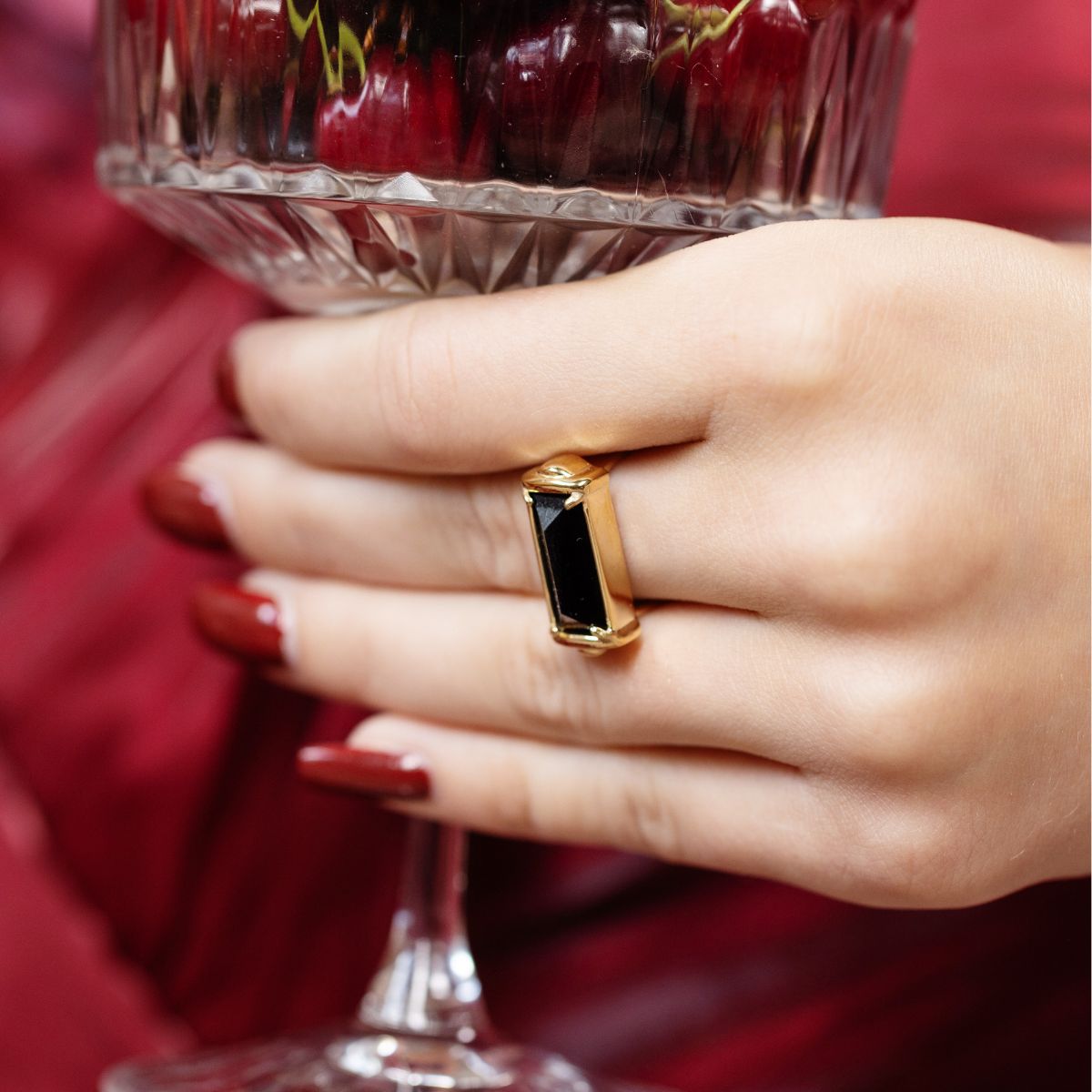 A close-up of a hand wearing a gold ring with a rectangular black stone, holding a clear glass filled with red cherries against a dark red fabric background.