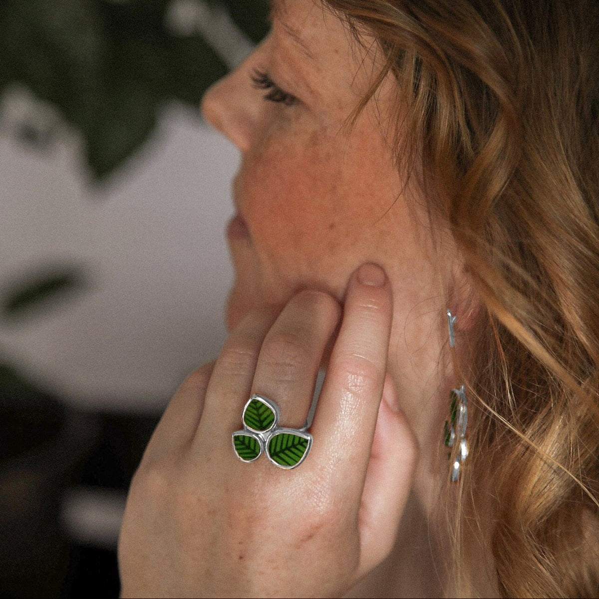Person wearing green leaf earrings with a plant in the background.