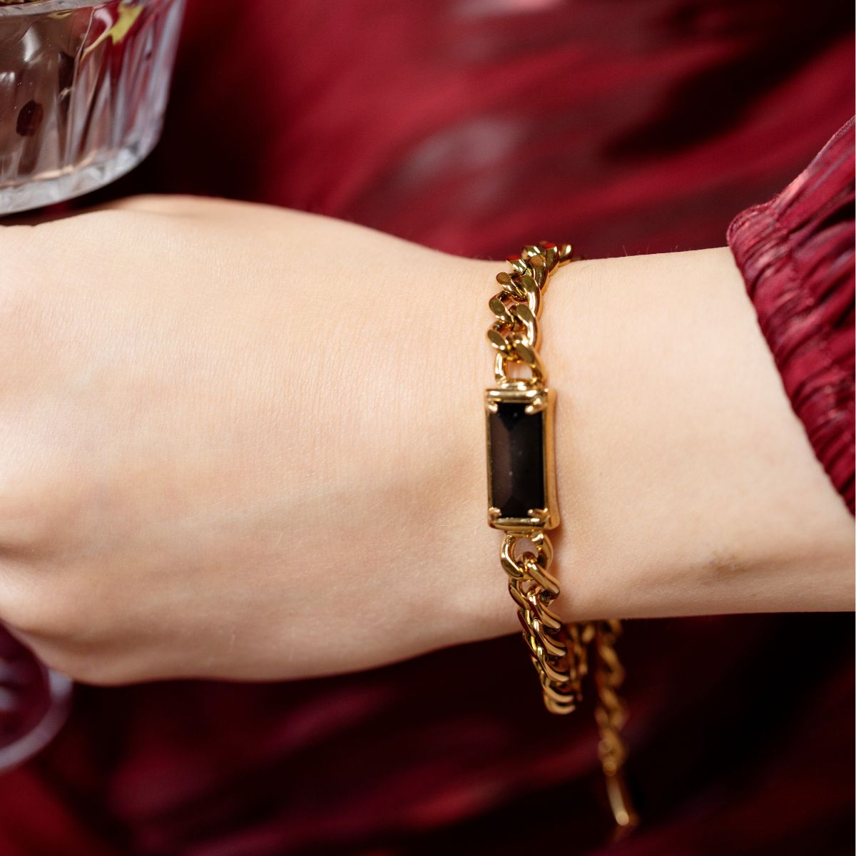 A gold-toned chain bracelet with a rectangular black inlay, shown worn on a person's wrist against a dark red fabric background.