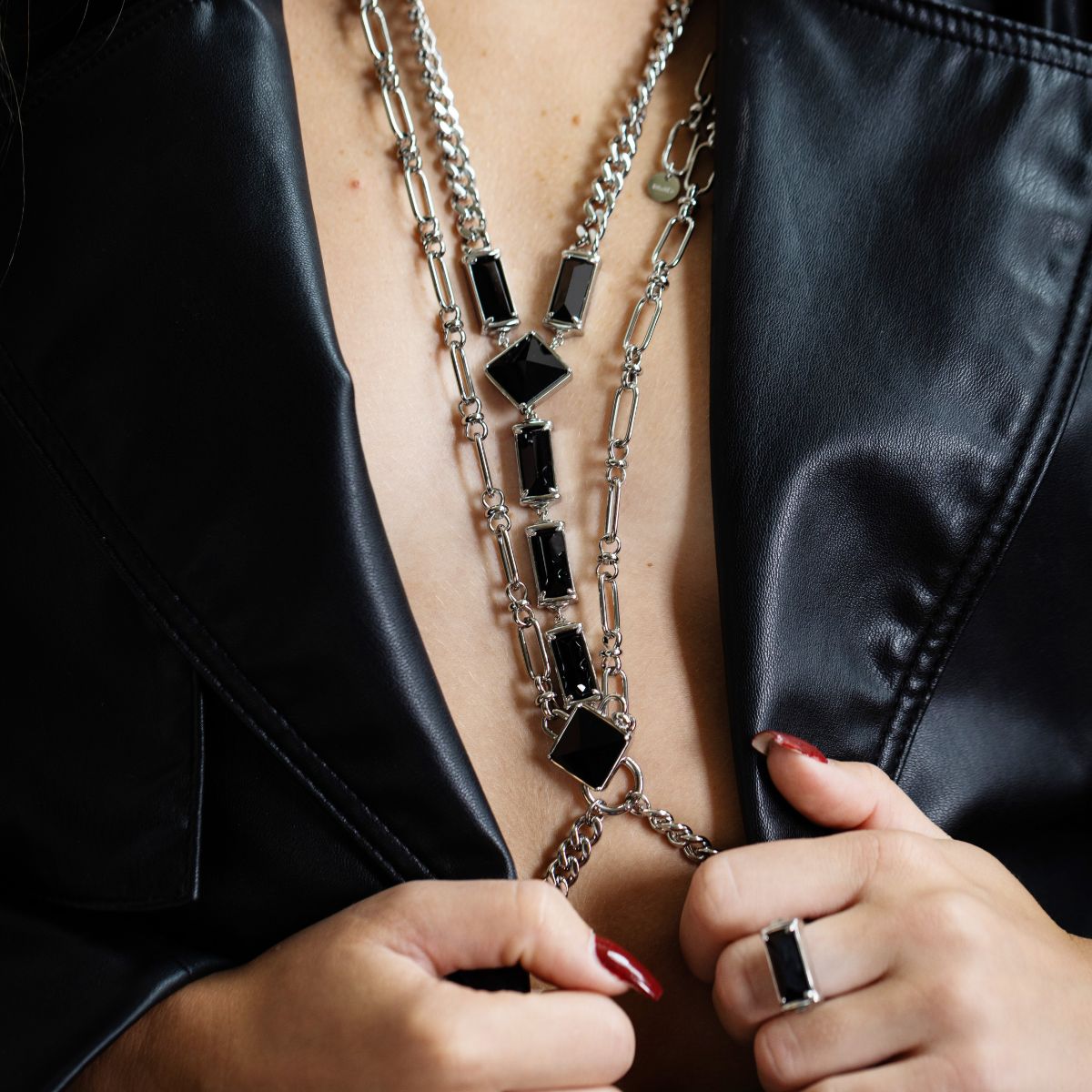 A close-up of a silver-toned double-strand necklace with rectangular and diamond-shaped black gemstones, worn by a model in a black leather jacket.
