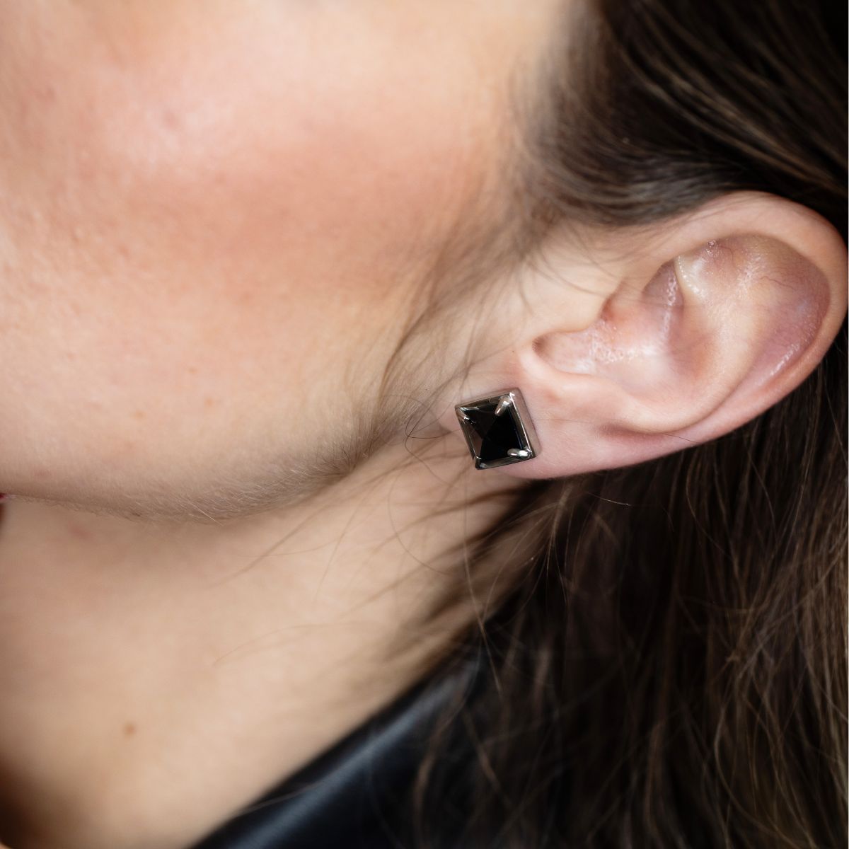 A close-up of a person's ear wearing a small square stud earring with a dark, polished rectangular gemstone in a silver-toned setting.