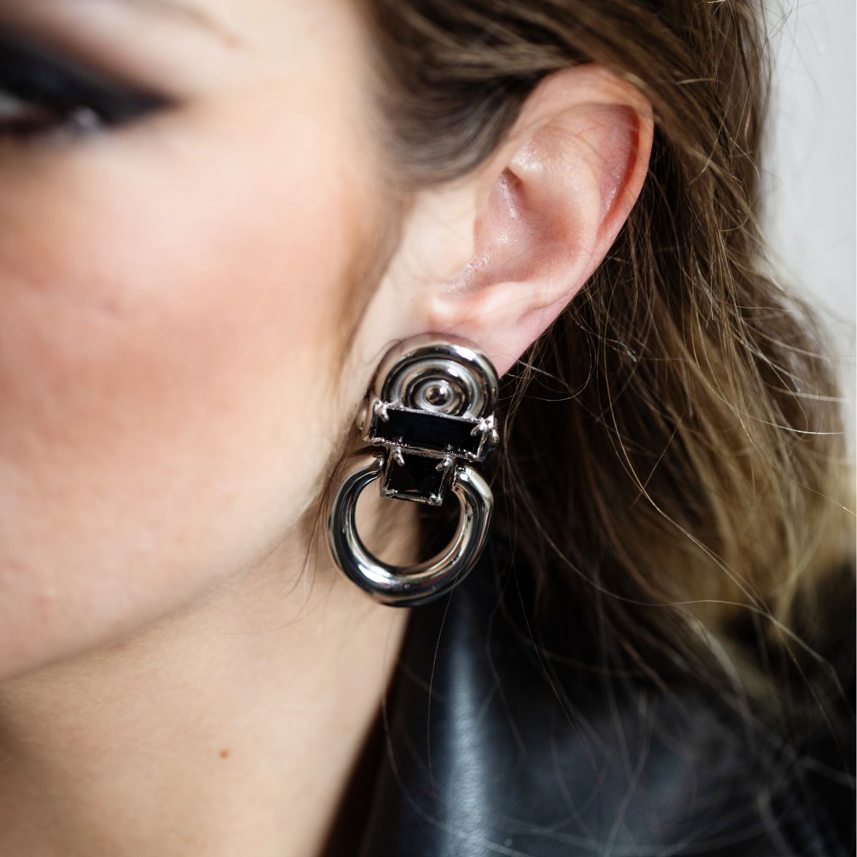 A close-up of a model's ear wearing a large, chunky silver-toned hoop earring with a black rectangular accent and a central spiral detail.