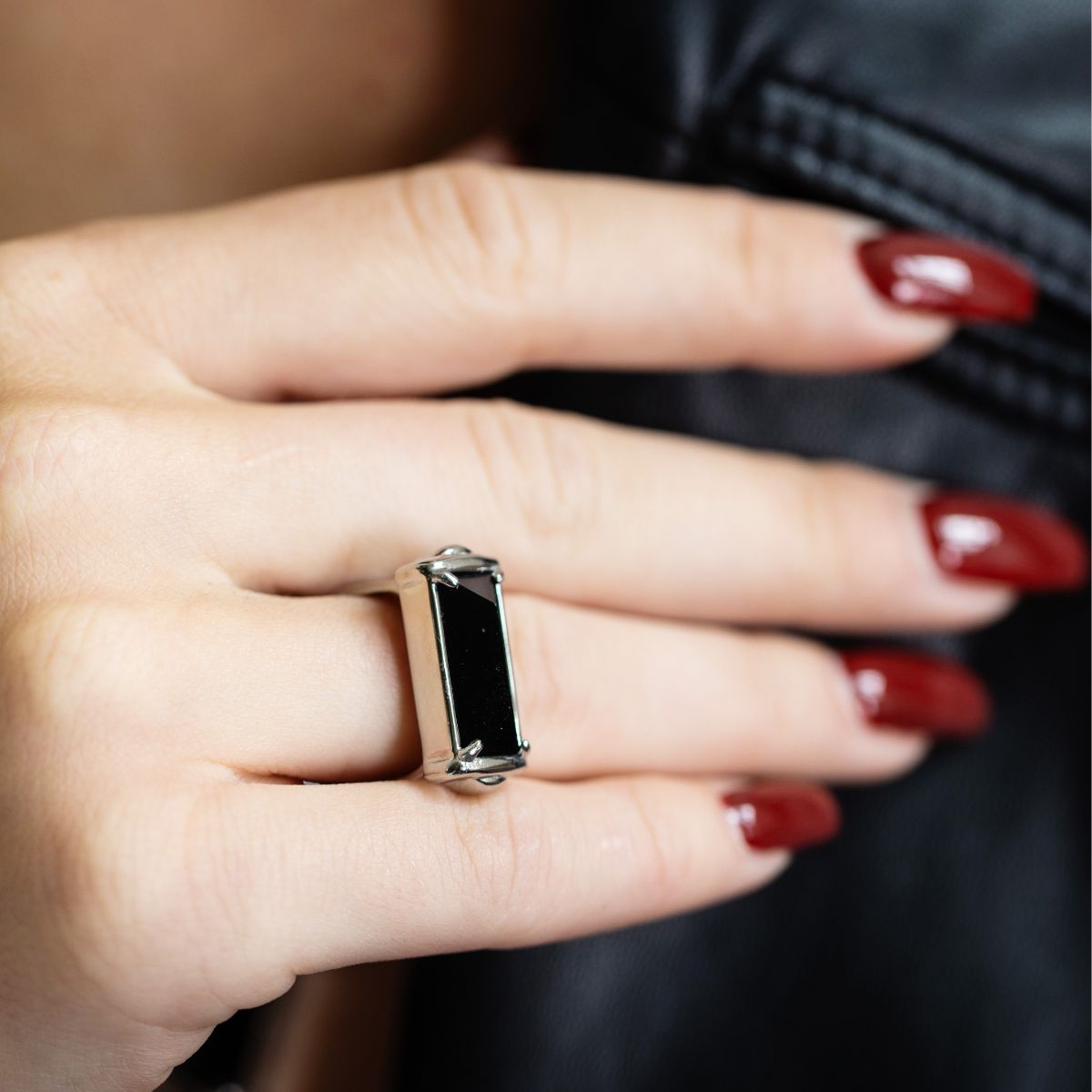 A silver-toned ring with a rectangular black inlay, worn on a hand with dark red manicured nails against a dark background.