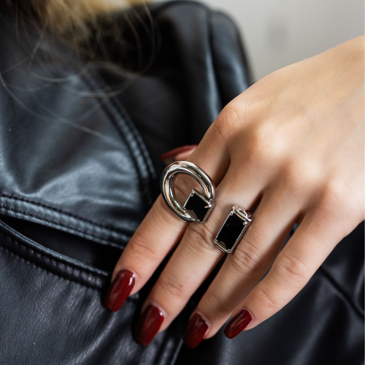 A close-up of a person's hand wearing two silver rings with black accents, resting against a black leather jacket. The hand has dark red manicured nails.