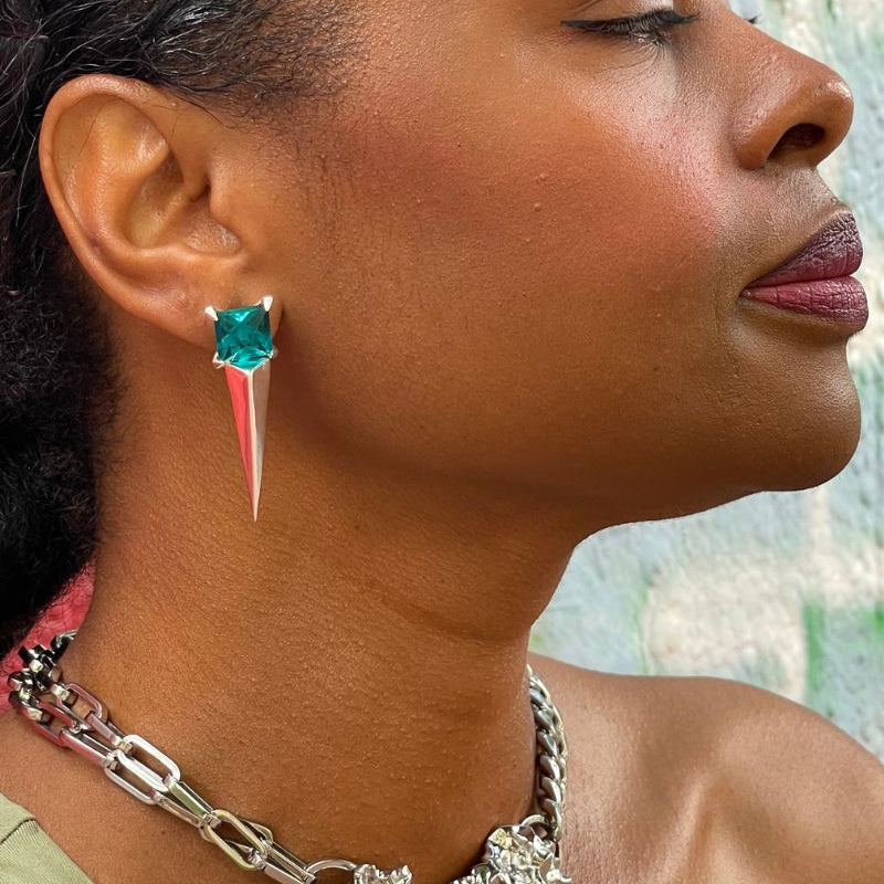 Close-up of a woman wearing a unique earring with a blue gem and red spike, set against a blurred natural background.