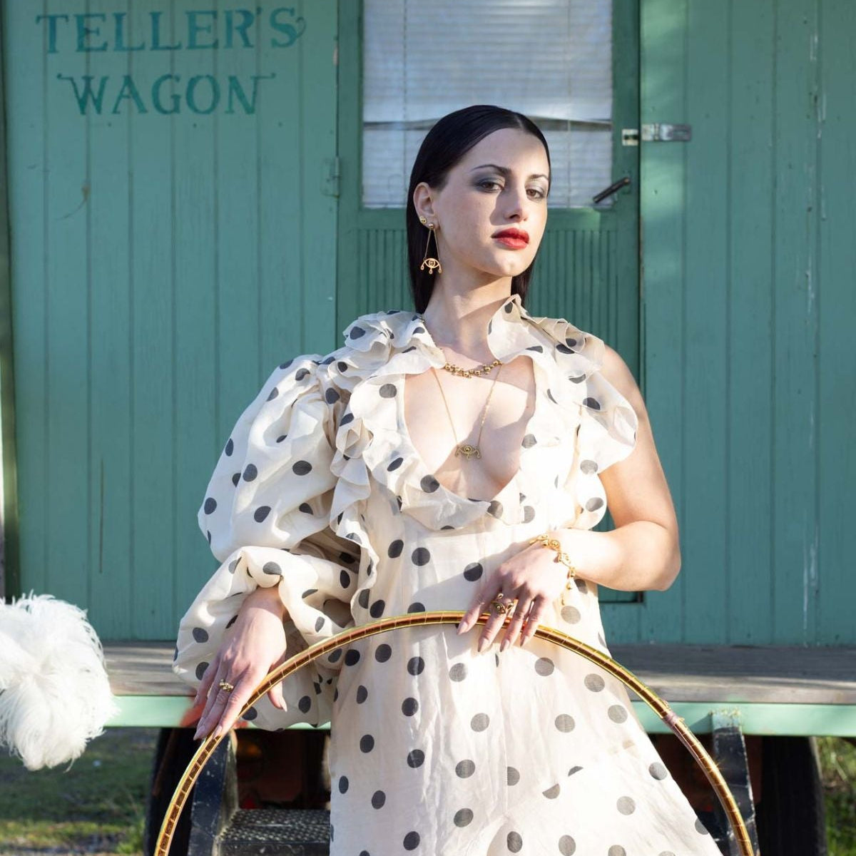 Woman in a white polka dot dress holding a hula hoop in front of a green building with text.