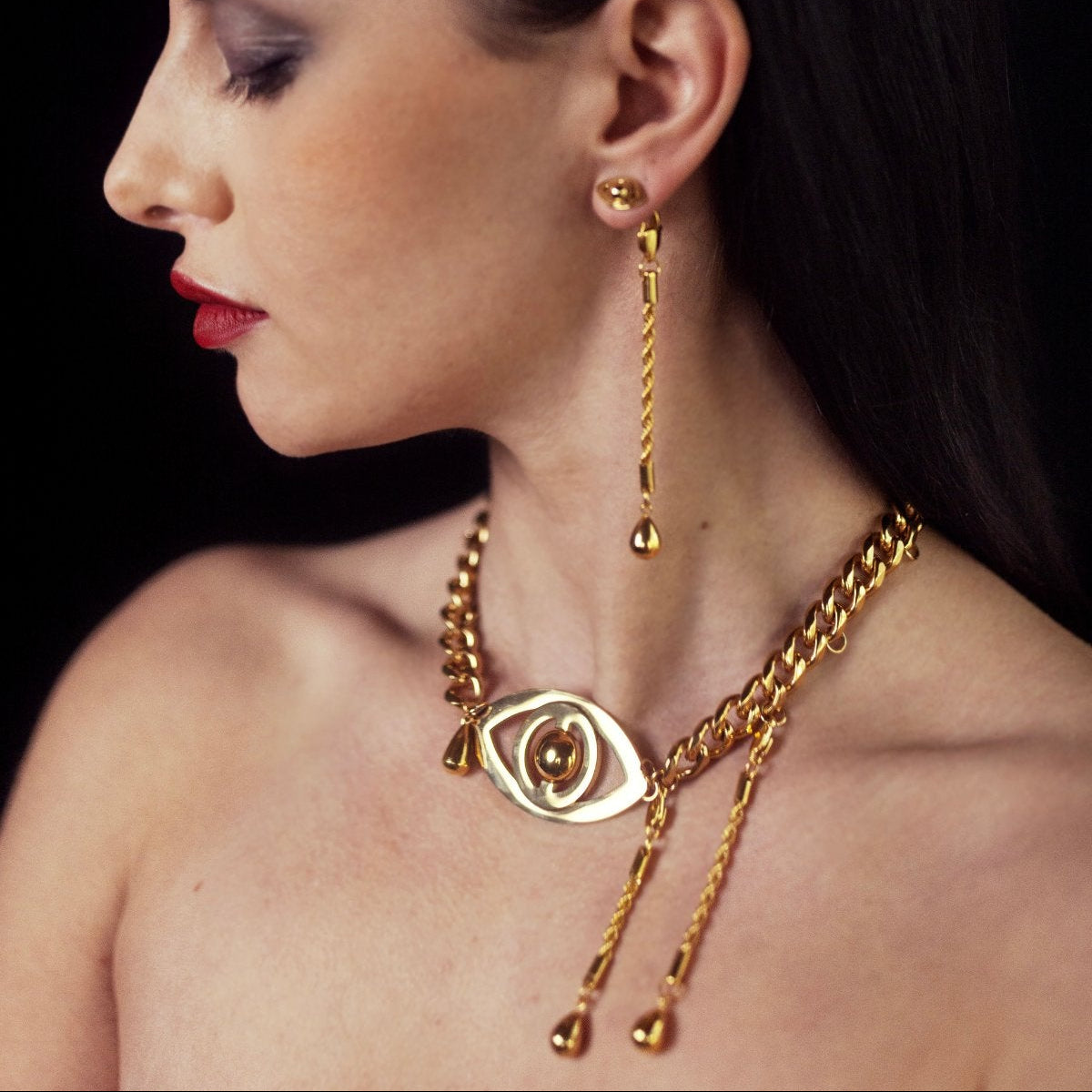 Woman wearing gold jewelry including an earring and necklace against a dark background