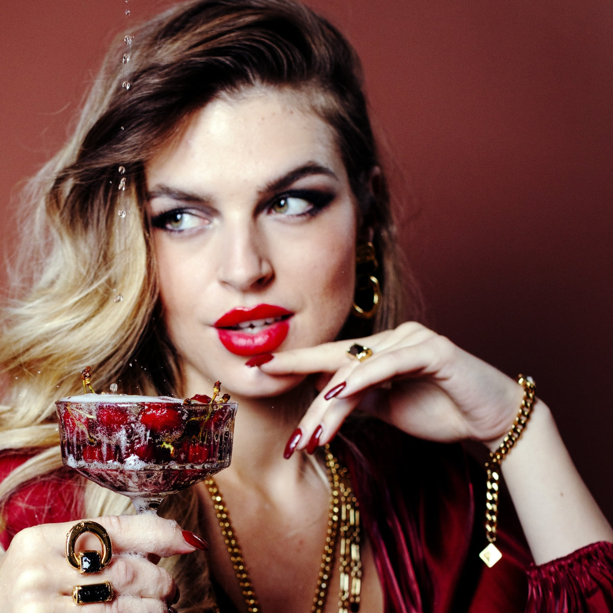 A woman with blonde-to-brown hair, wearing a red velvet top and gold jewelry, holding a glass filled with red liquid and strawberries. Water droplets splash near the glass against a dark reddish-brown background.