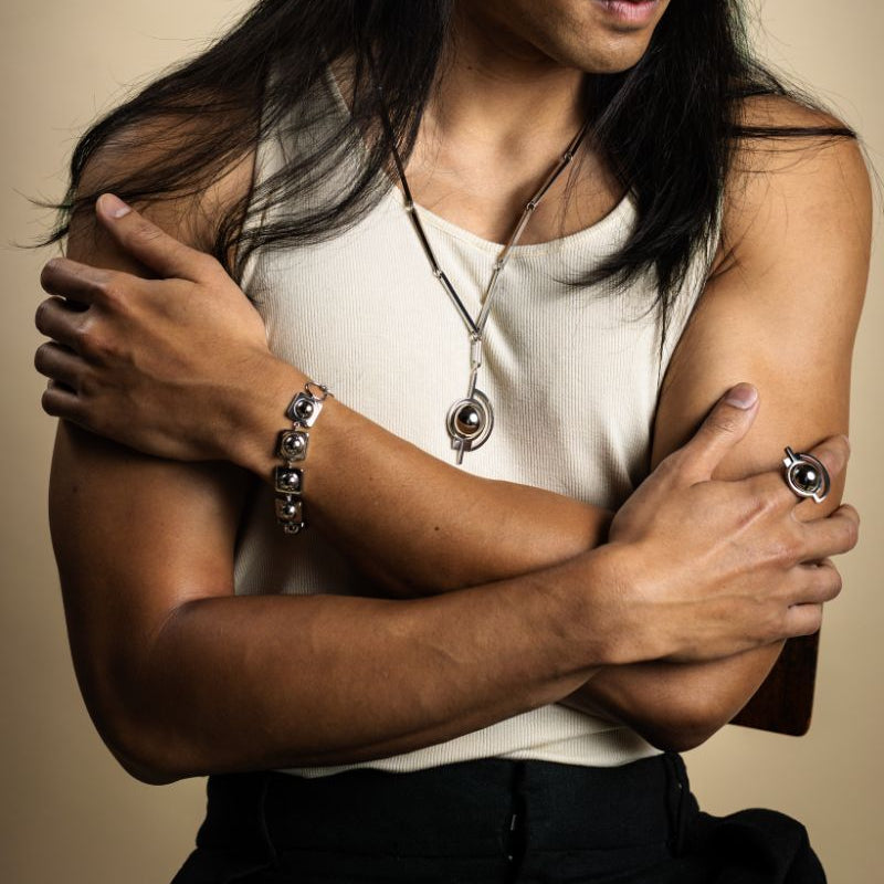 Man with long hair wearing a necklace, ring, and bracelet against a beige background